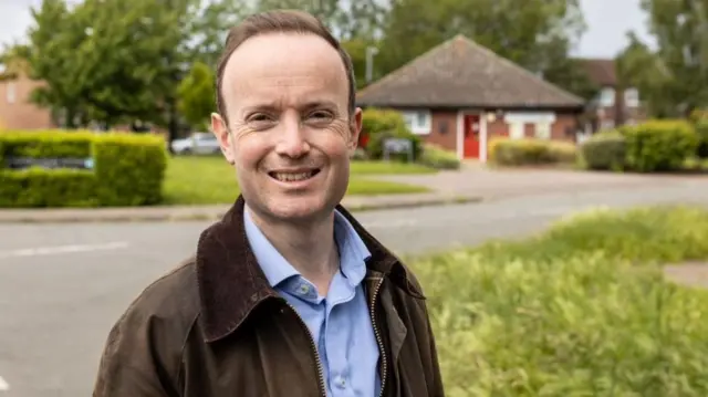 Richard Rout wearing a blue shirt and a brown jacket standing on the side of a road