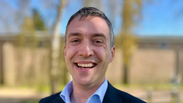 Adrian Ramsay smiles at the camera. He wears a navy suit jacket and light blue shirt underneath.
