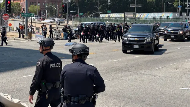 Dozens of police walk in a line across the road in sunny LA, next to two police cars