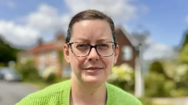 Anne-Marie Robb smiles at the camera while standing outside. She has dark hair that is tied back behind her head and wears glasses with a bright green jumper.