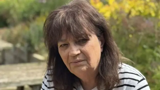 Jenny Kirtley smiles at the camera while sitting on a park bench outside. She has shoulder length dark hair and wears a white and black striped top.