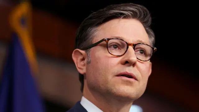 Mike Johnson looks onwards past the camera. He has brown hair and brown eyes. He is wearing a white shirt and navy blazer. He is wearing brown-rimmed glasses.