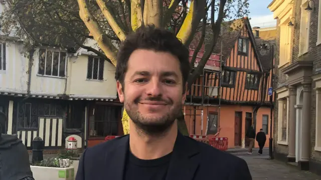 Jack Abbott smiles at the camera standing outside in a town. He has dark hair and wears a black suit jacket with a black T-shirt. There are Tudor/medieval style buildings behind him in Ipswich town centre