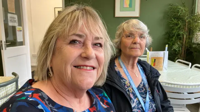 Nicky Corbett (left) and Lesley Hill (right) sit next to each other in a cafe. They are smiling at the camera.