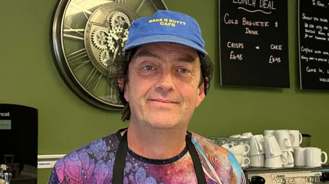 Barry Smith smiles at the camera in a cafe. He wears a blue cap, has dark hair and wears a multi-coloured top with a black apron on.