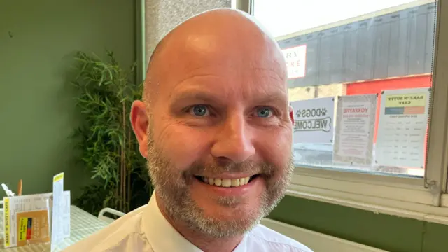 Tom Pawle smiles at the camera while sitting in a cafe. He is bald and has a beard. He wears a white shirt and tie.