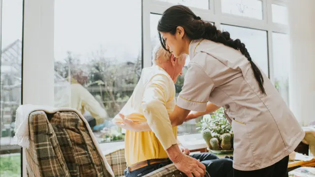 A young female carer helps an elderly male to get dressed by pulling on his jumper, as he sits in an armchair