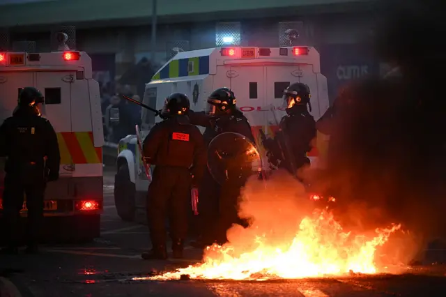 Police stand beside a landrover in riot gear. There is a fire beside them