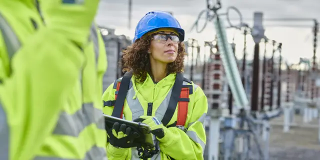 A construction worker stands on site with a pen and pad, and inspects the site with a hard hat