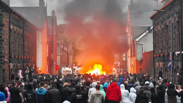 A large group of people standing while a fire burns at the top of a street