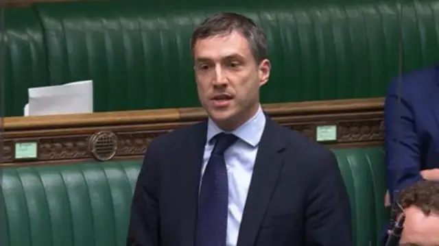 Headshot of Ramsay who wears a navy suit stood up talking in front of green leather benches
