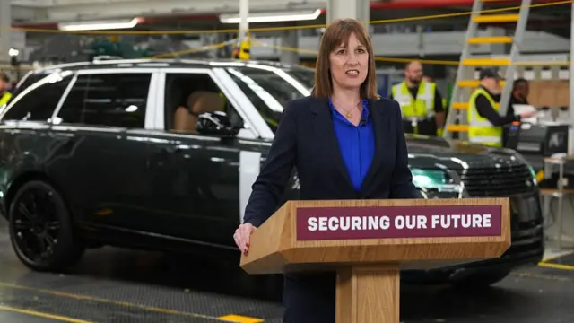 Rachel Reeves, standing at a lectern with the sign 'securing our future' on the front, delivers a speech in front of a Range Rover at a car factory