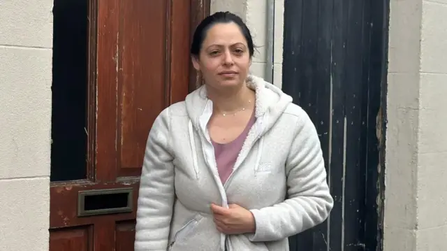 A woman standing in front of a damaged front door of a house. She has dark hair pulled back into a pony tail and is wearing a grey hooded top, with a pink top underneath it.