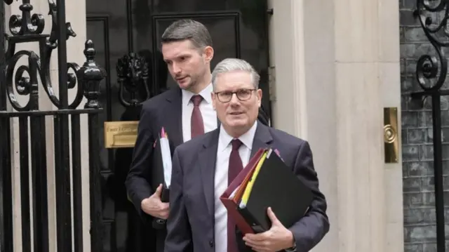 Starmer leaving No.10 Downing Street, wearing suit with folders in hand.