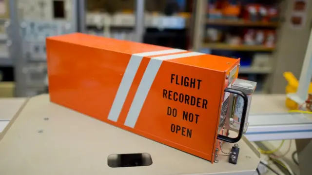 A bright orange, rectangular metal box with the words 'flight recorder do not open' in black on the side, sits on a workbench