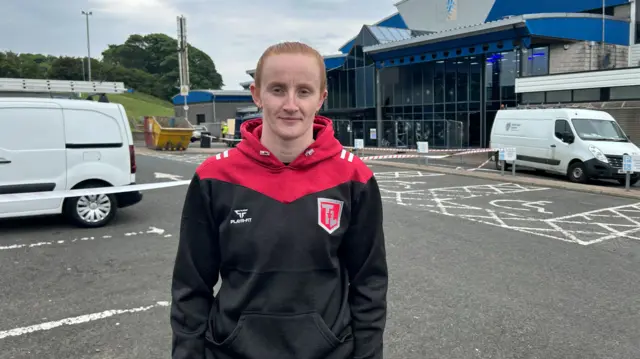 Danielle Hill, she has red hair and is wearing a black and red jumper. She is standing in front of Larne Leisure Centre