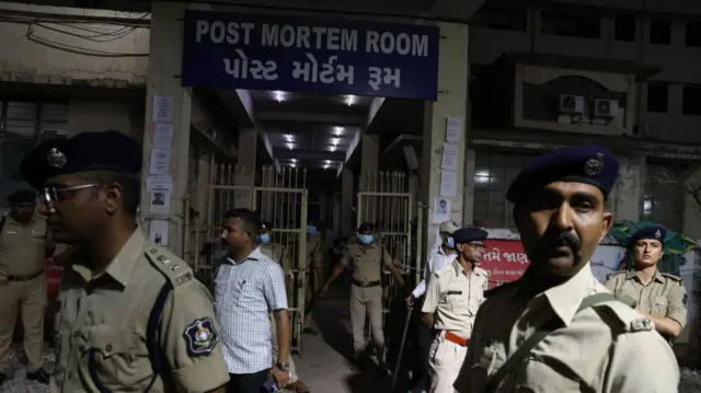 Indian military personnel in green and beige uniform and dark berets stand outside the post mortem room (as indicated by a blue sign and white writings on top of the main gated-entrance) of a civil hospital in Ahmedabad