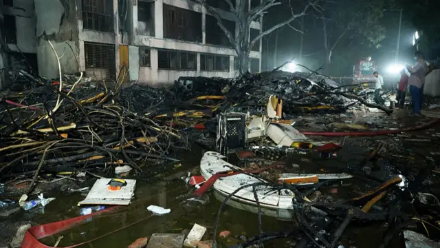 Overnight view of Ahmedabad crash site. Debris strewn over the ground illuminated by large lights as some rescue workers stand to the far right