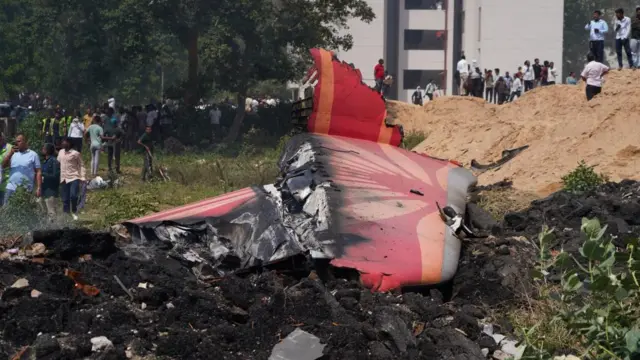 Tail fin of plane on the ground, debris strewn around as people stand nearby.