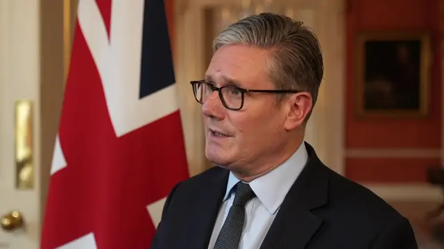 Mid-shot of Sir Keir Starmer in black suit, light blue shirt and dark tie as he speaks insider Number 10, a Union flag to his right