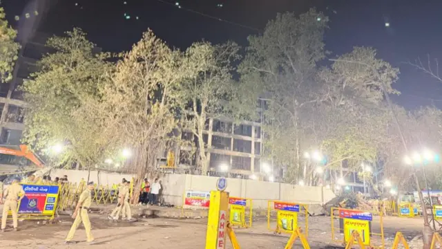 Night shots of plane crash site in Ahmedabad illuminated by large industrial lights. A damaged building in the background with some trees around it