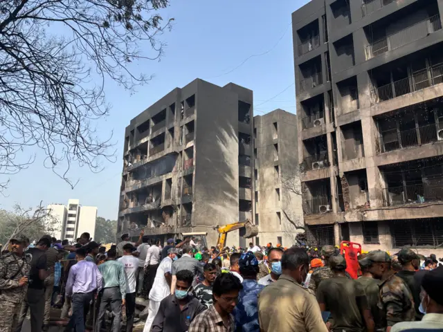 A crowd of people and a digger at work near a tower block which has been damaged in crash