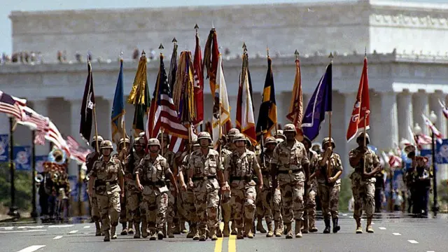 A group of military personnel in khaki fatigues march down a wide street carrying various flags
