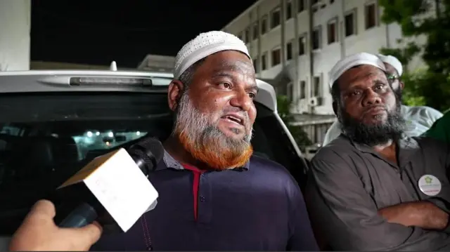 Sameer Shaikh stands leaning against the back of a white car as he speaks to the press. He's wearing a dark blue, long-sleeved polo shirt, a long beard with a ginger tip and a white hat. To his left is a man also leaning against the car in a matching white hat and grey, open-collared shirt