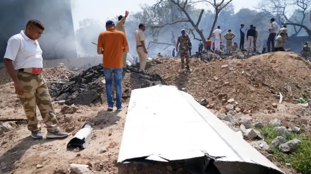 Part of plane on floor, debris around with people gathered and smoke rising behind.