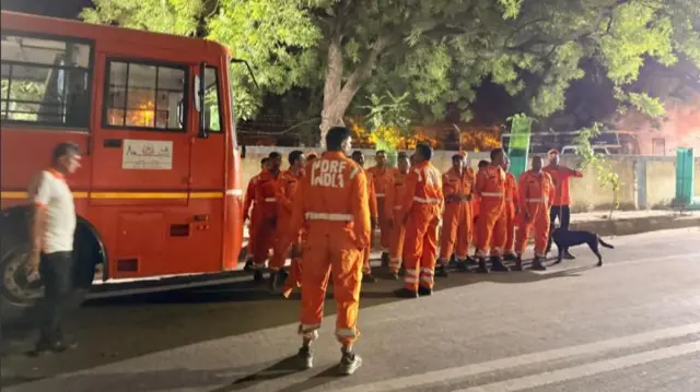 Members of India's National Disaster Response Force in their orange uniforms stand on the streets next to an orange bus outside the crash site