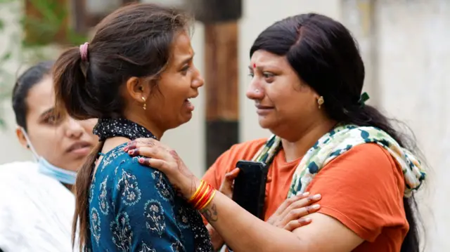 Two women hold each other and cry. Another woman looks on from behind them, concerned.