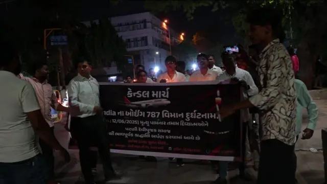 Men hold a sign while walking through a street in Ahmedabad at night.