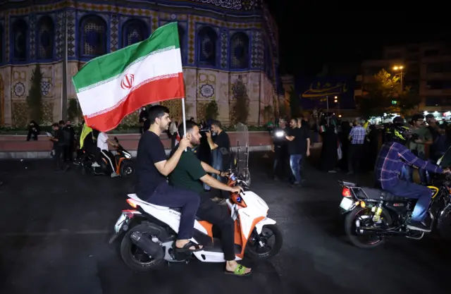 Two men are on a motorcycle, with the one in back hold an Iranian flag.