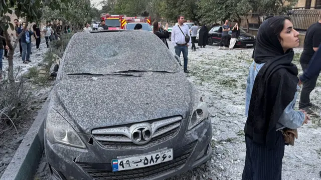 A woman with a black headscarf standing in front of a black sedan covered in ash