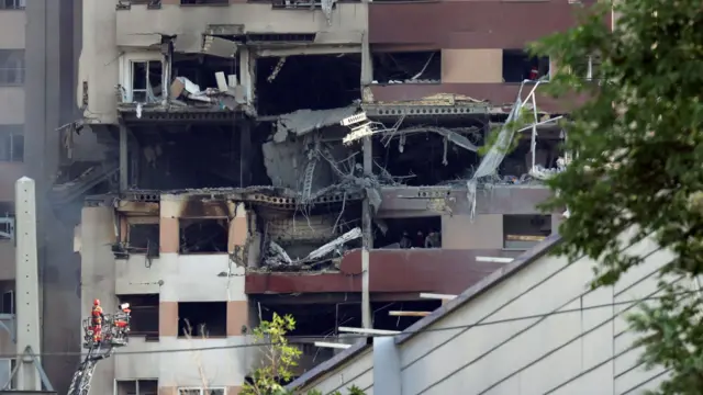 A building stands hollowed out with two emergency service personnel in high-vis on a metal structure looking towards it
