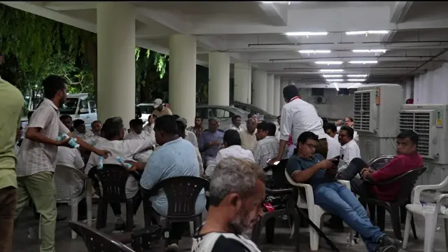 A group of people at the Civil Hospital in Ahmedabad sit on black and white plastic chairs at night.