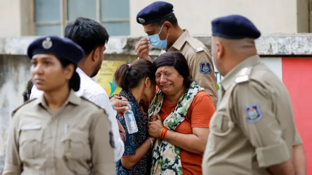 Two women with long brown hair hug each other while crying. Four officials stand nearby.