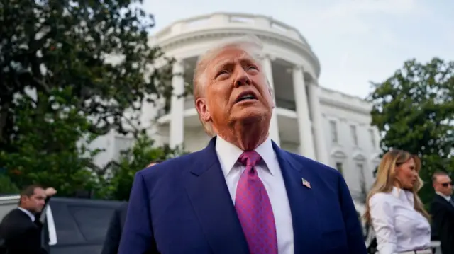 Mid-shot of Donald Trump standing outside the White House in a dark blue suit, white shirt and pink and purple patterned tie. The White House main building is behind him, to his left is his wife Melania Trump in a white shirt and skirt talking, blurred. Secret Service agent stands behind Trump with a hand on a black car