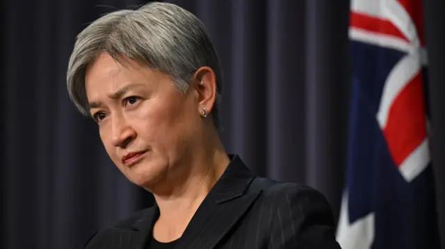 Penny Wong looks sternly, standing in front of a blue curtain and an Australian flag