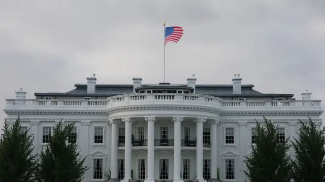 Outside shot of the White House at dusk, the American flag flying from a pole on top of the roof