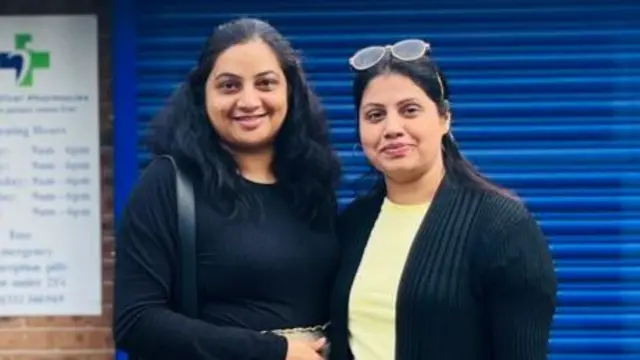 Trupti Telrandhe and Kalyani Brahmbhatt stand in front of a pharmacy and are smiling at the camera. Both have black hair and brown eyes. Trupti is wearing a black, long-sleeved top. Kalyani is wearing a yellow t-shirt and black cardigan, and she has sunglasses placed on top of her head.