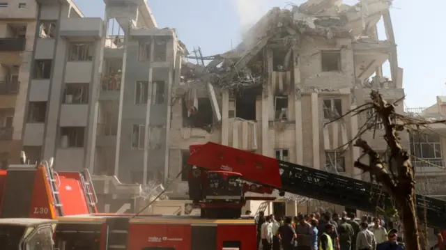 A building stands with the top part collapsed into rubble. A red truck is in the foreground with people standing looking up at it