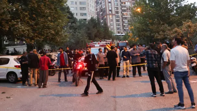 People gather on a Tehran street. There is an emergency vehicle in the background.