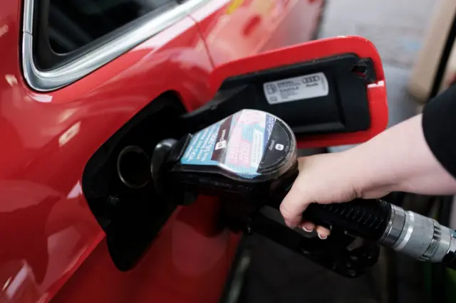 A woman holds a petrol pump in her right hand as she refuels a red car.