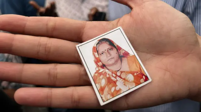 A passport size photo of Sarla Thakor wearing an printed orange sari, seen in the palm of a hand