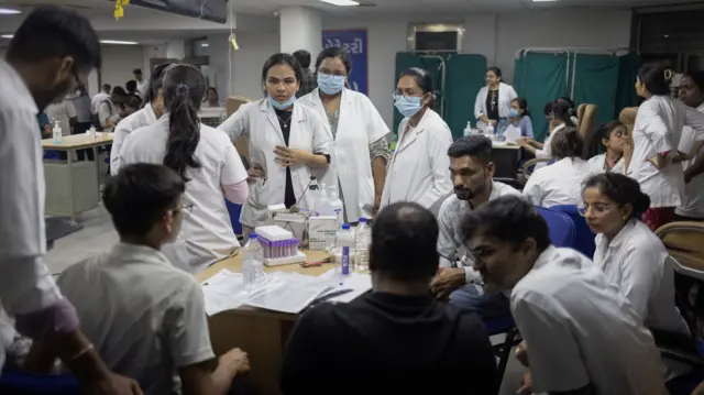 A group of men and women in lab coats sit around a table that holds water bottles and clear plastic test tubes with purple lids.