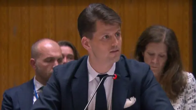 A man speaking into a small mic as he looks off to one side. Some people are sat behind him
