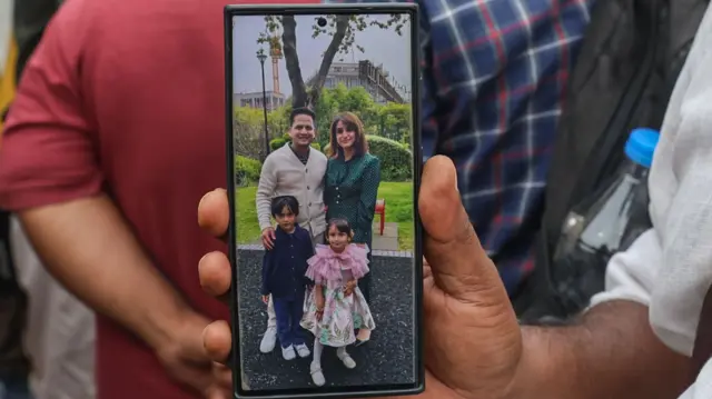 A man's hand holds up a phone. On the screen are a man and woman, smiling, and standing with a young boy and girl.
