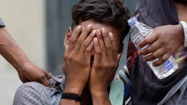 A man holds his hands up to his eyes as he sobs. Two other people's hands rest on his shoulders, comforting him. One person, whose face is not in the shot, holds a water bottle.