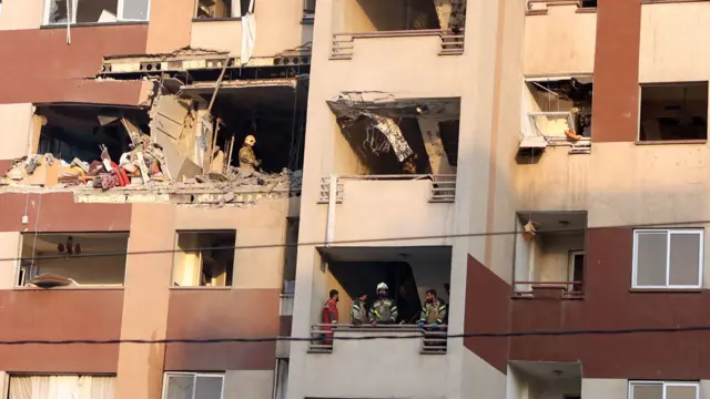 A shot of what looks like a residential building. Rubble can be seen in various windows and firefighters stand inside the building